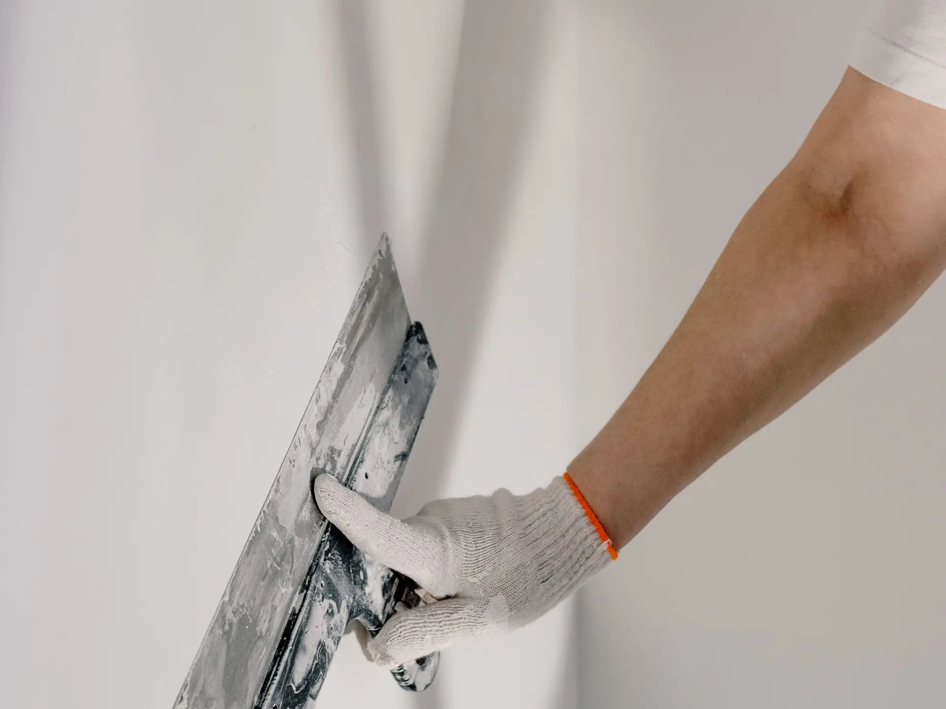 Main gantée qui passe un large couteau à enduire sur un mur blanc
