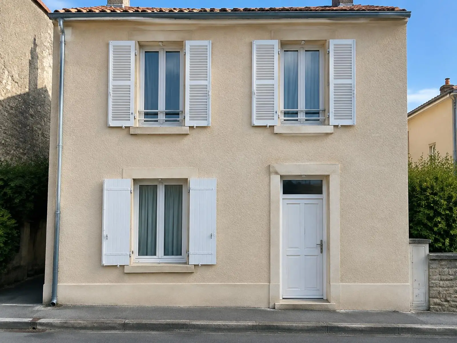 La même façade au crépi beige propre et uni, volets persiennés et porte blancs, trottoir au pied du mur