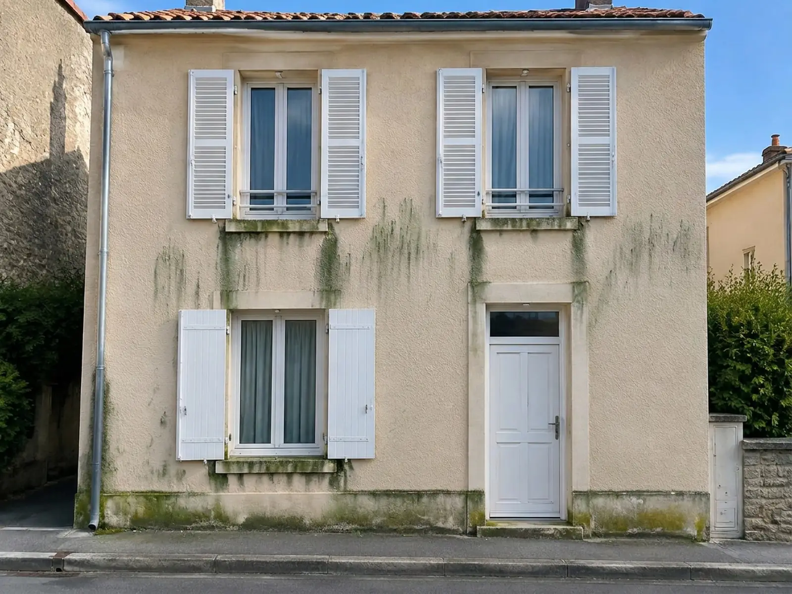 Façade sur rue au crépi beige marquée de traînées vertes sous les appuis et au pied du mur, volets blancs