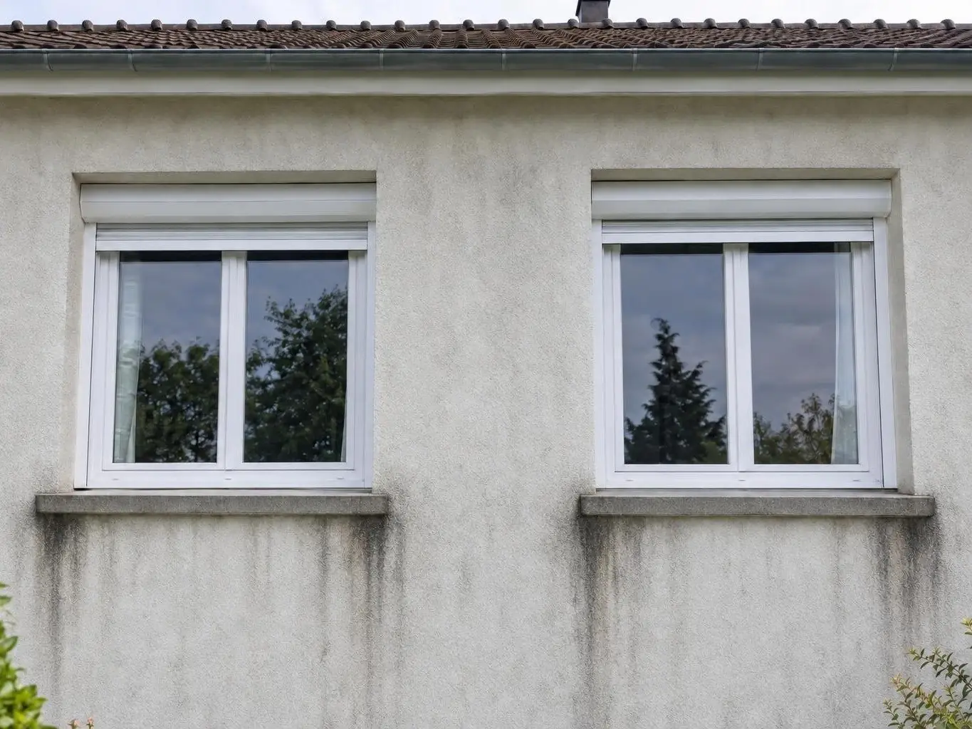 Façade en crépi blanc cassé grisé, longues traînées noires sous les appuis de deux fenêtres blanches à volet roulant