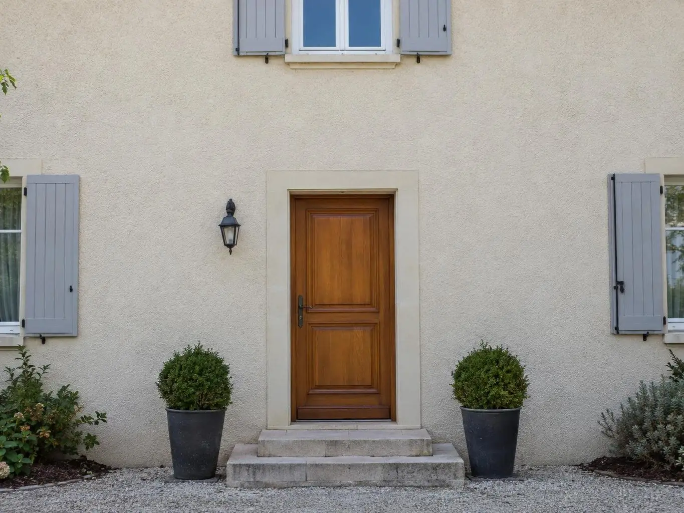 Façade en crépi beige uniforme au-dessus d’une porte d’entrée en bois verni, applique noire et deux buis en pot