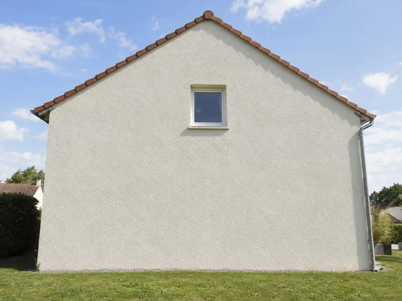 Pignon de pavillon en crépi gratté blanc cassé neuf et uniforme sous un toit de tuiles rouges, pelouse au pied du mur