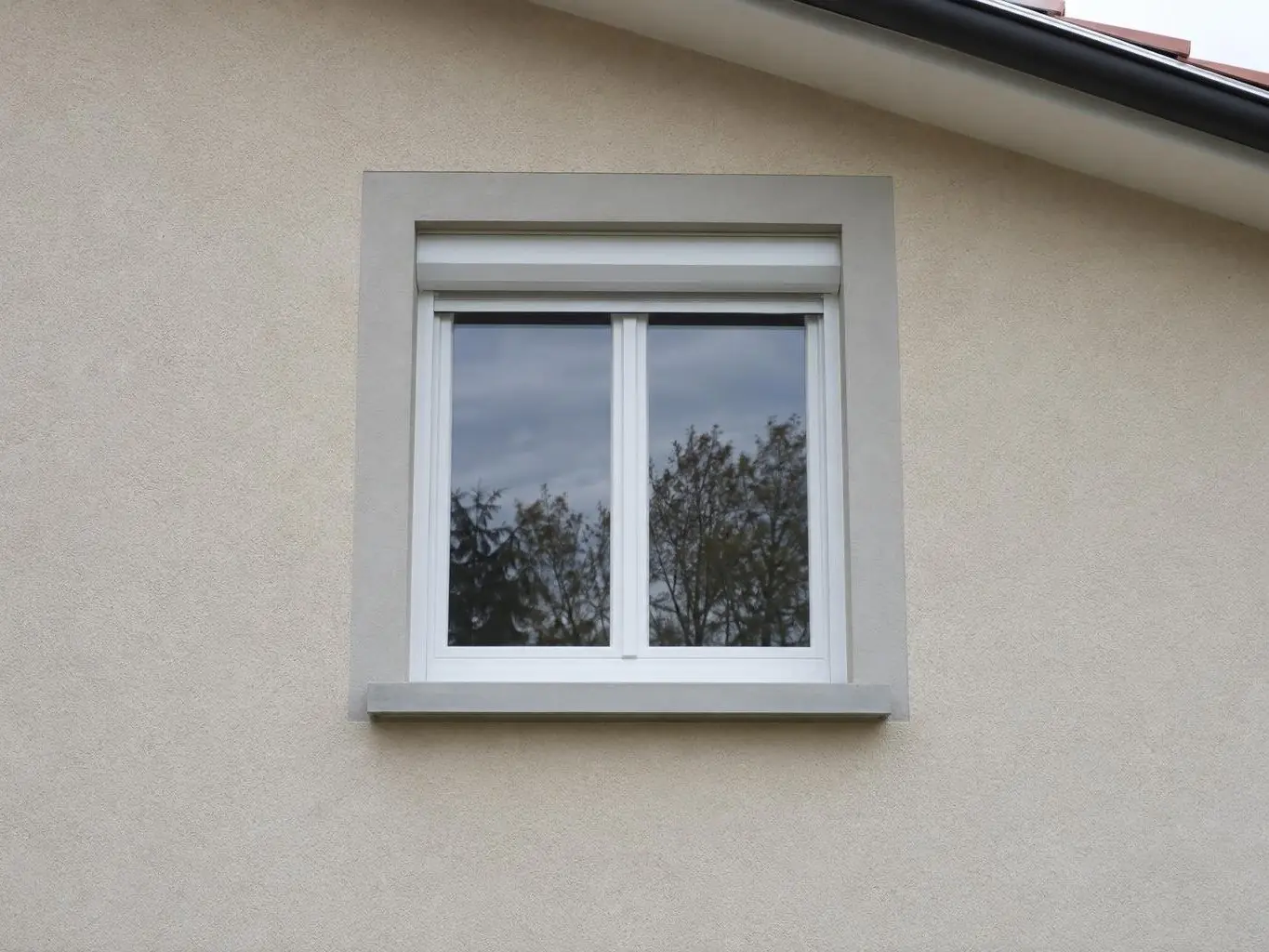 Fenêtre blanche à volet roulant encadrée d’une bande d’enduit gris clair, appui en béton lisse aux arêtes nettes