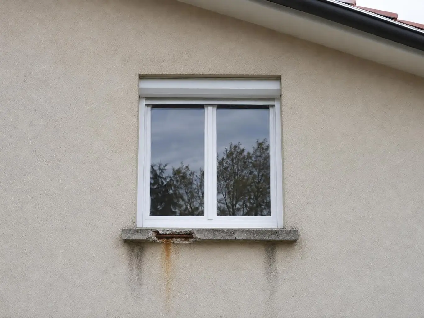 Fenêtre blanche à volet roulant au-dessus d’un appui en béton éclaté, ferraille rouillée et coulures brunes sur le crépi