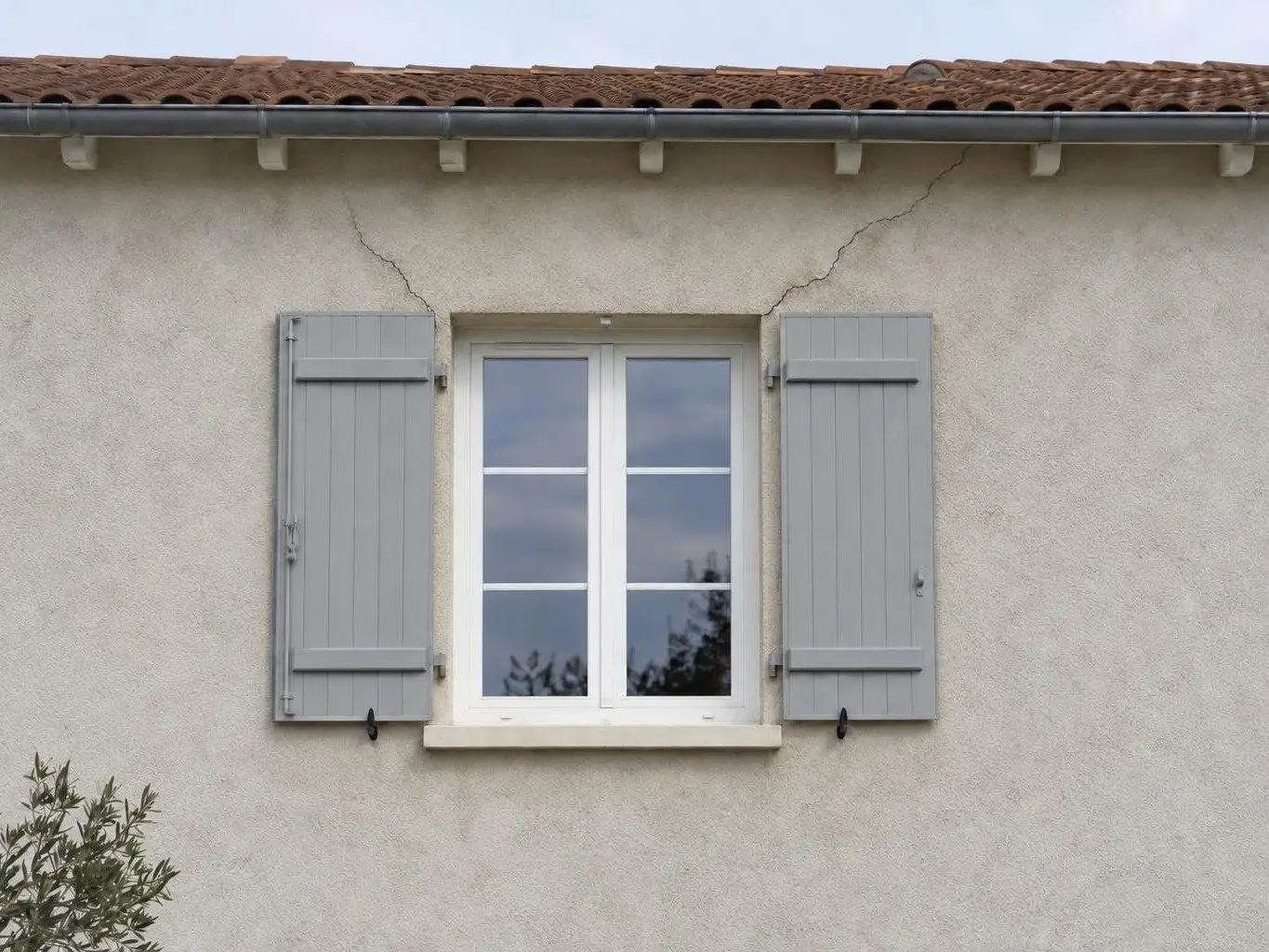 Fenêtre blanche à volets gris clair dans un crépi blanc cassé, deux fissures en biais partant de ses coins vers le toit