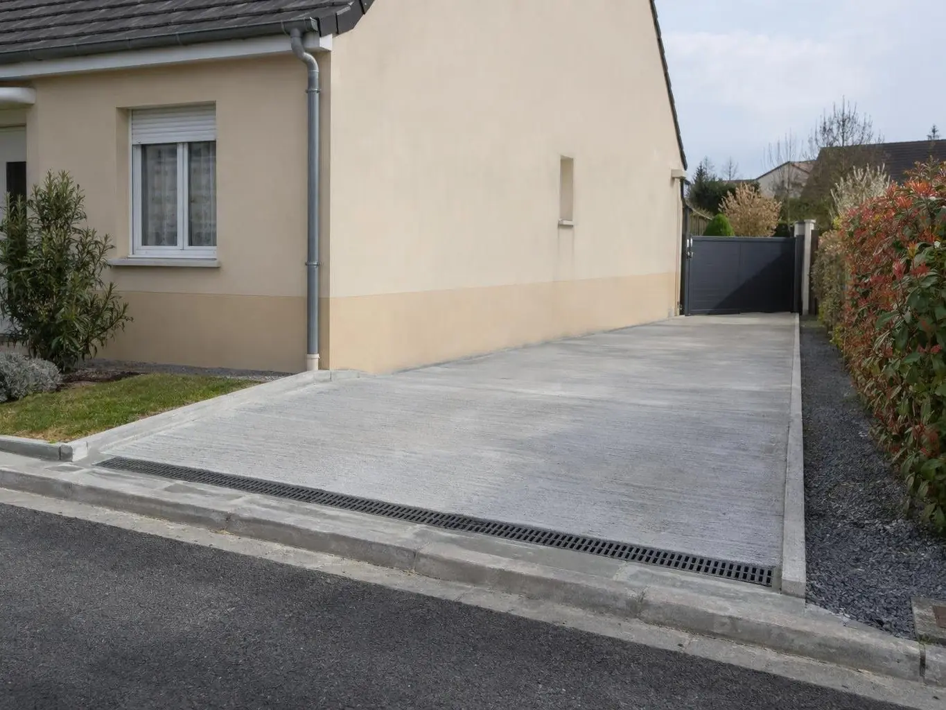 Place de parking en béton balayé gris clair le long d’un pavillon beige, grille d’évacuation au bord du trottoir
