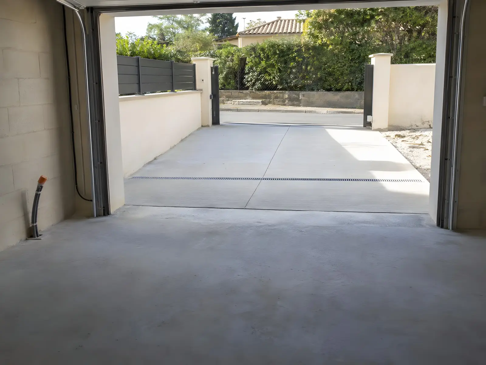 Intérieur d’un garage neuf vide : sol en béton, grande porte ouverte sur une allée bétonnée et un portail