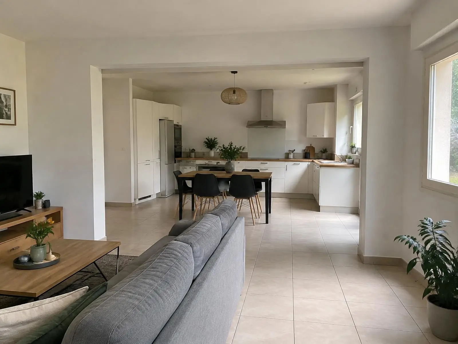 Séjour ouvert en grand sur la cuisine sous une poutre peinte en blanc, même carrelage beige d’une pièce à l’autre
