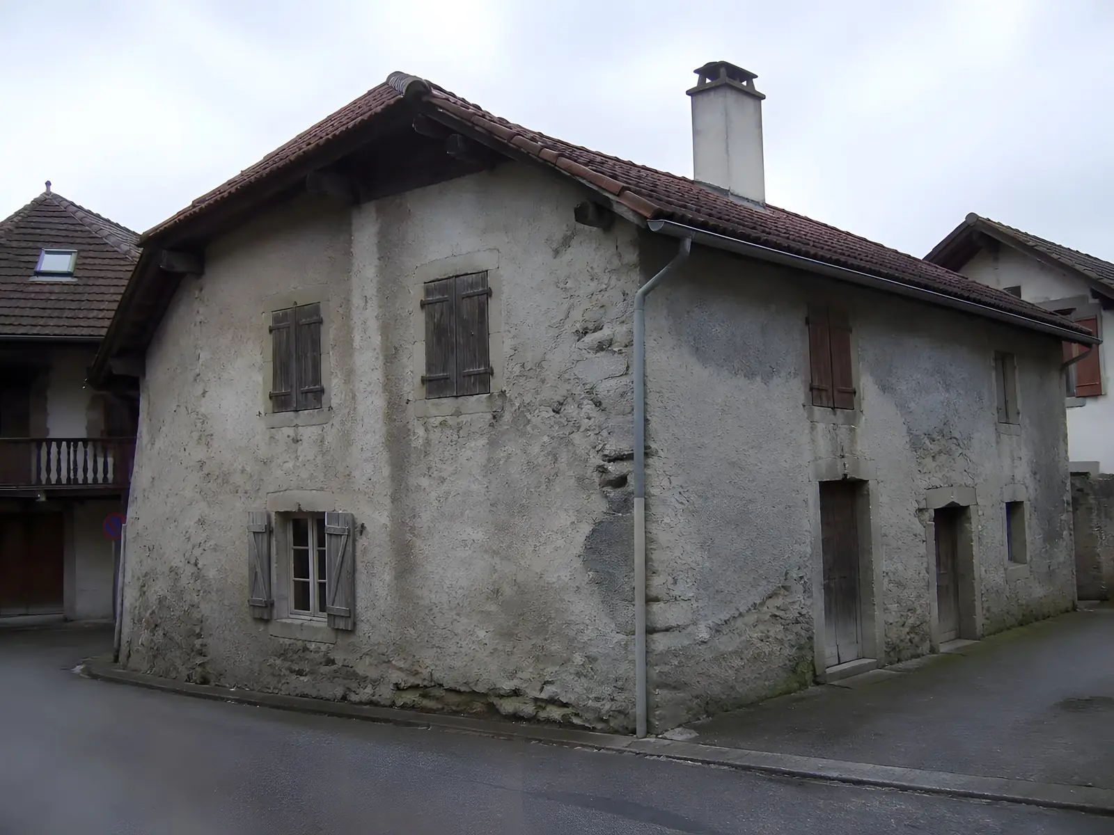 Maison de village ancienne à l’enduit gris encrassé, décollé par plaques, volets en bois délavés