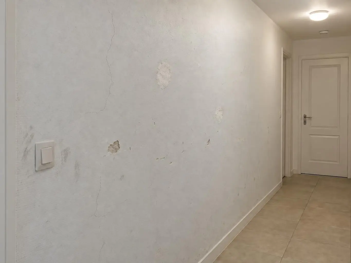 Mur de couloir peint en blanc grisé, fissures fines, éclats et rebouchages plus clairs autour de l’interrupteur