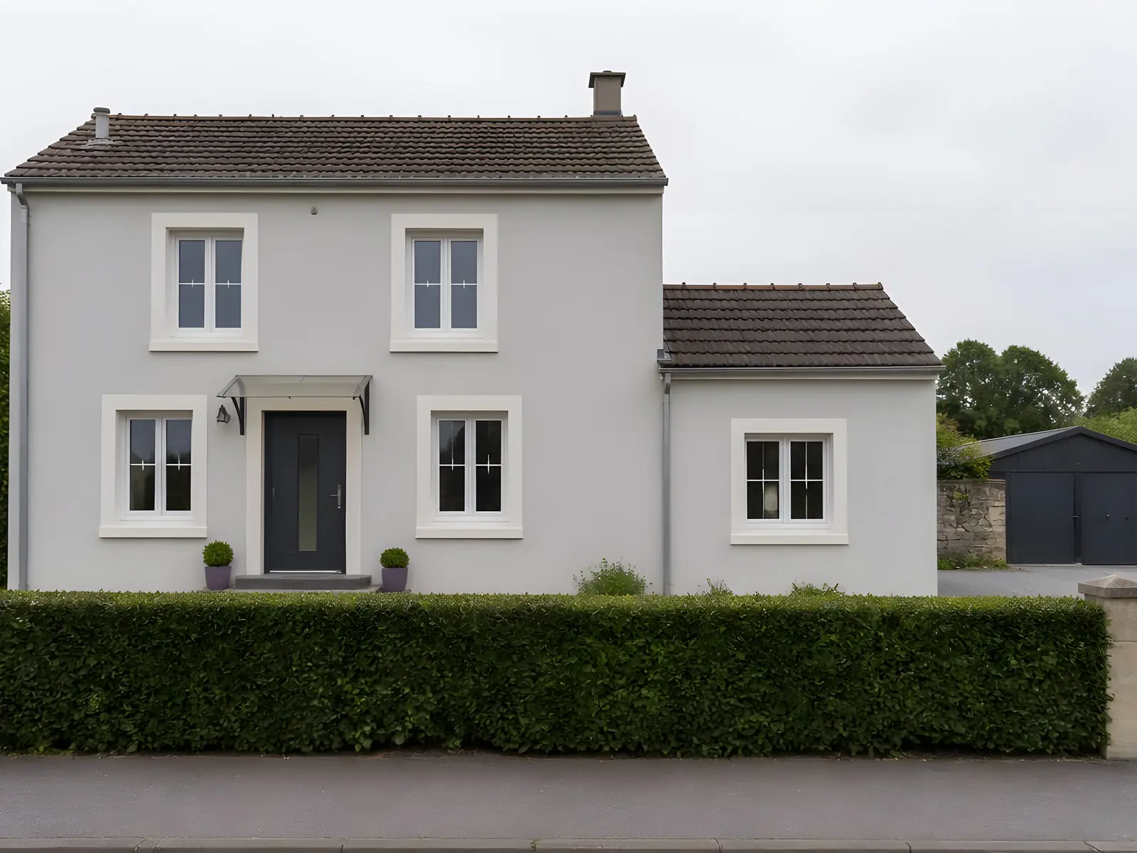 Le même pavillon repeint en gris clair, tours de fenêtres blanc cassé et porte anthracite, derrière sa haie