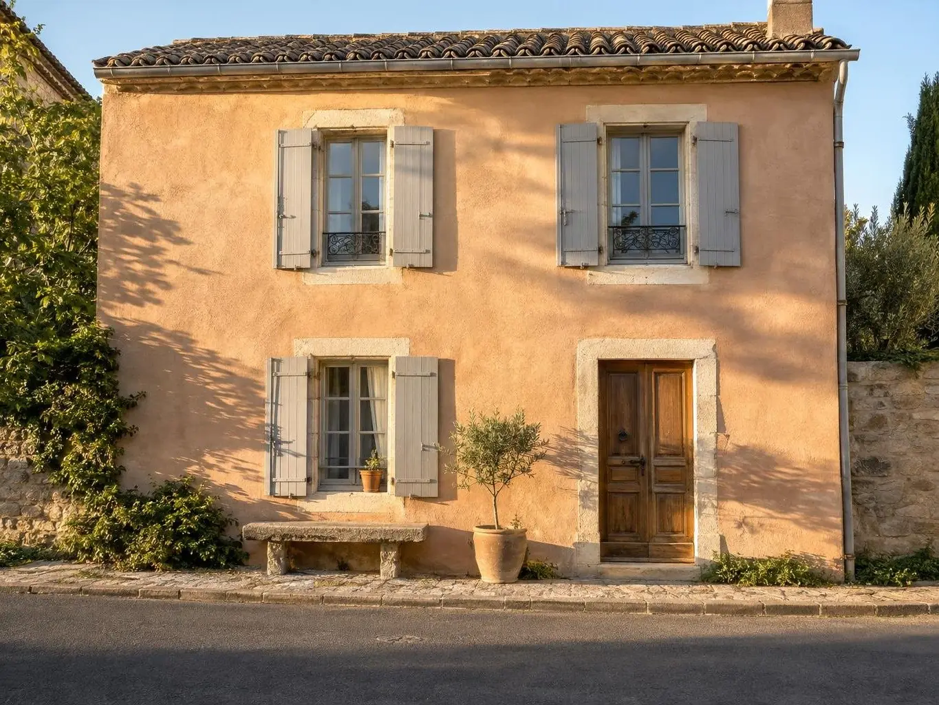 Façade de maison de village peinte à la chaux ton sable rosé, encadrements en pierre claire, volets gris, banc de pierre