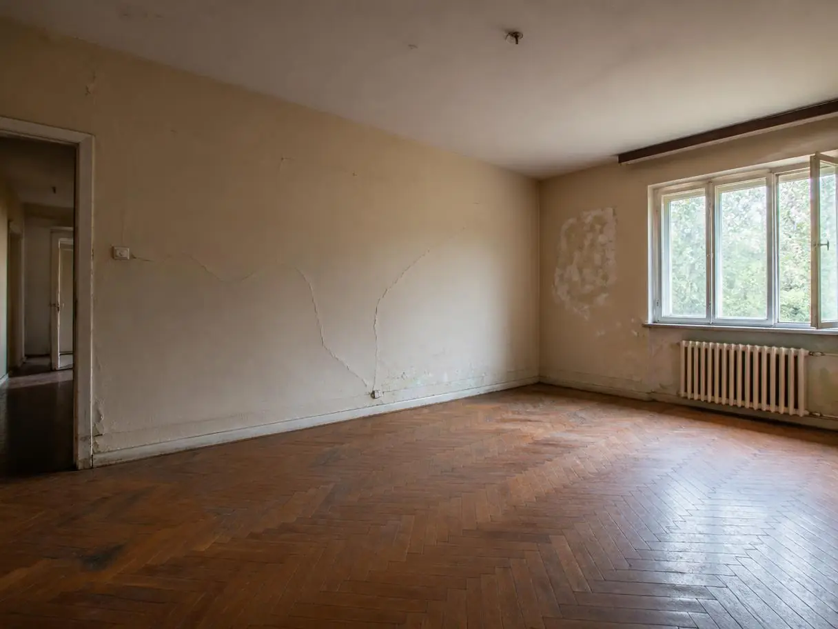 Séjour vide aux murs beiges fissurés, plâtre écaillé près de la fenêtre et du radiateur, avant les travaux