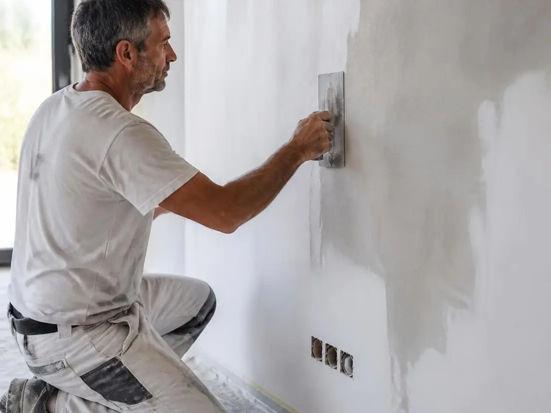 Peintre à genoux sur un sol bâché lissant l’enduit sur un mur blanc avec une lisseuse en inox