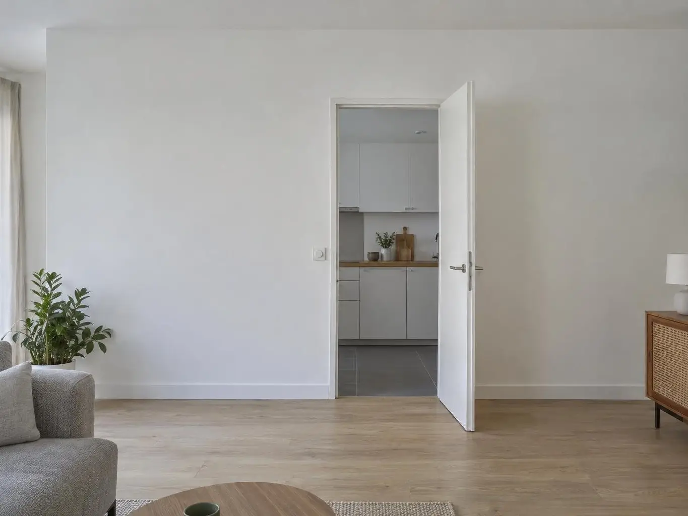 Séjour au parquet clair fermé par une cloison blanche, porte ouverte sur la cuisine, interrupteur près du chambranle