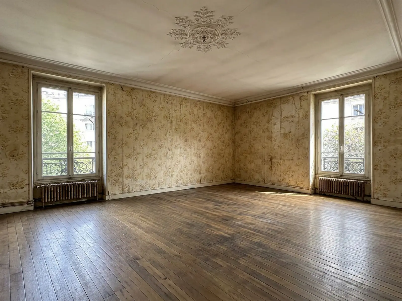 Grande pièce ancienne au papier peint jauni, deux hautes fenêtres à garde-corps et vieux parquet