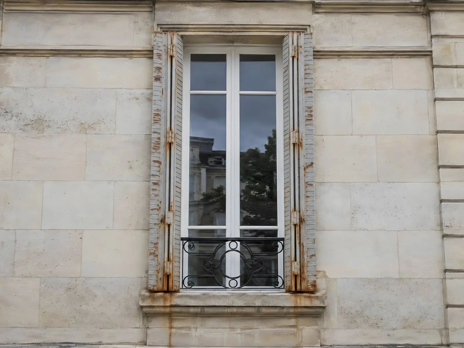 Persiennes métalliques grises écaillées et piquées de rouille, traînées orange sur la pierre sous la fenêtre