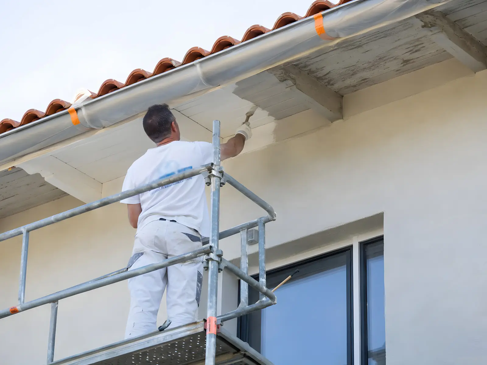 Peintre debout sur un échafaudage qui repeint en blanc au pinceau le dessous de toit d’une maison aux tuiles rondes